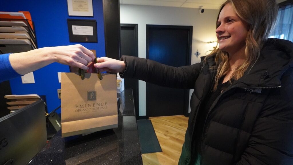 Happy customer receiving a paper bag filled with professional skincare products from Uptown Haven, an ideal complement to a wellness gift card.