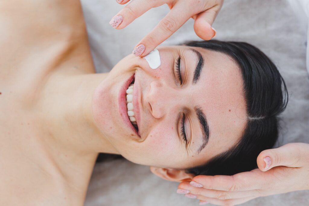 The face of a young woman to whom a beautician applies cream to her skin.