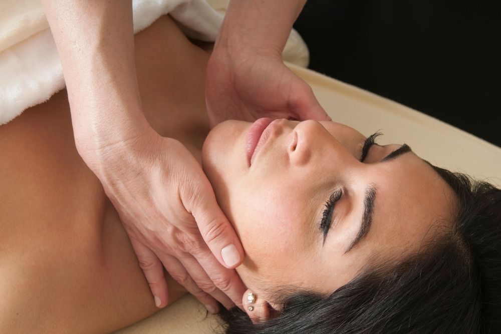A therapist gently massaging the jawline and neck of a relaxed woman lying face up during a facial lymphatic drainage session.