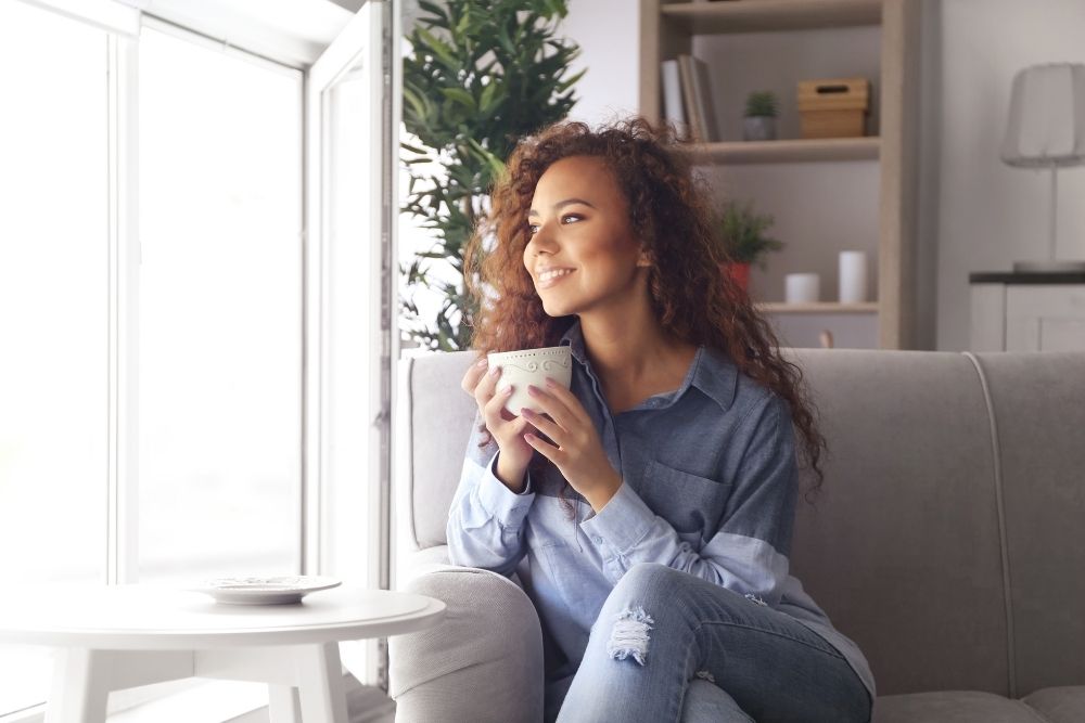 A relaxed woman with curly hair sitting on a couch, smiling and holding a warm mug near a bright window, representing wellness and peace.
