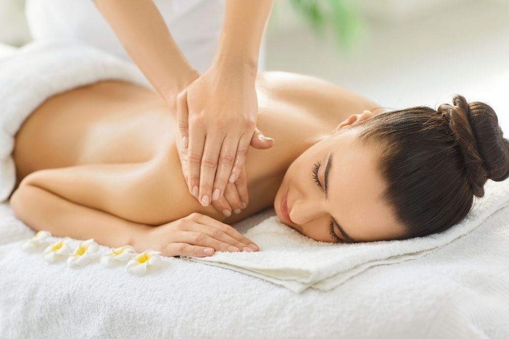 A woman lying face down on a massage table with white flowers nearby, smiling serenely as a therapist gently works on her shoulder.
