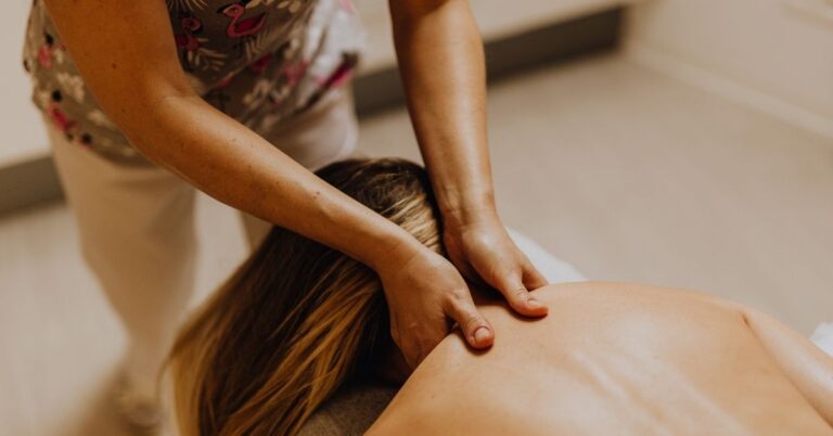 Close-up of a massage therapist applying gentle pressure to a woman's upper back and shoulder area in a warm, dimly lit spa setting.