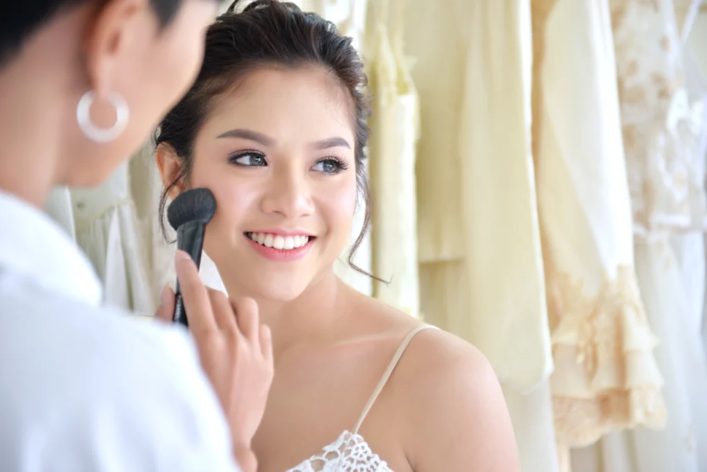 Smiling bride-to-be having makeup applied with a brush in front of a rack of wedding gowns