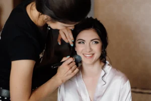 Makeup artist applying foundation with a brush to a smiling bride in a silk robe on her wedding morning